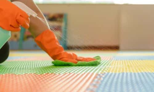 foto di una mano con guanto arancione che con un panno verde sanifica con il giusto prodotto la superficie dei giochi dei bambini in un asilo